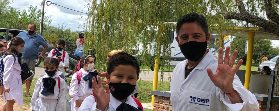 Darío Greni y un alumno de la escuela, vestidos con túnica, saludan mirando a la cámara. Detrás se ven otros estudiantes. Se encuentran fuera de la escuela.