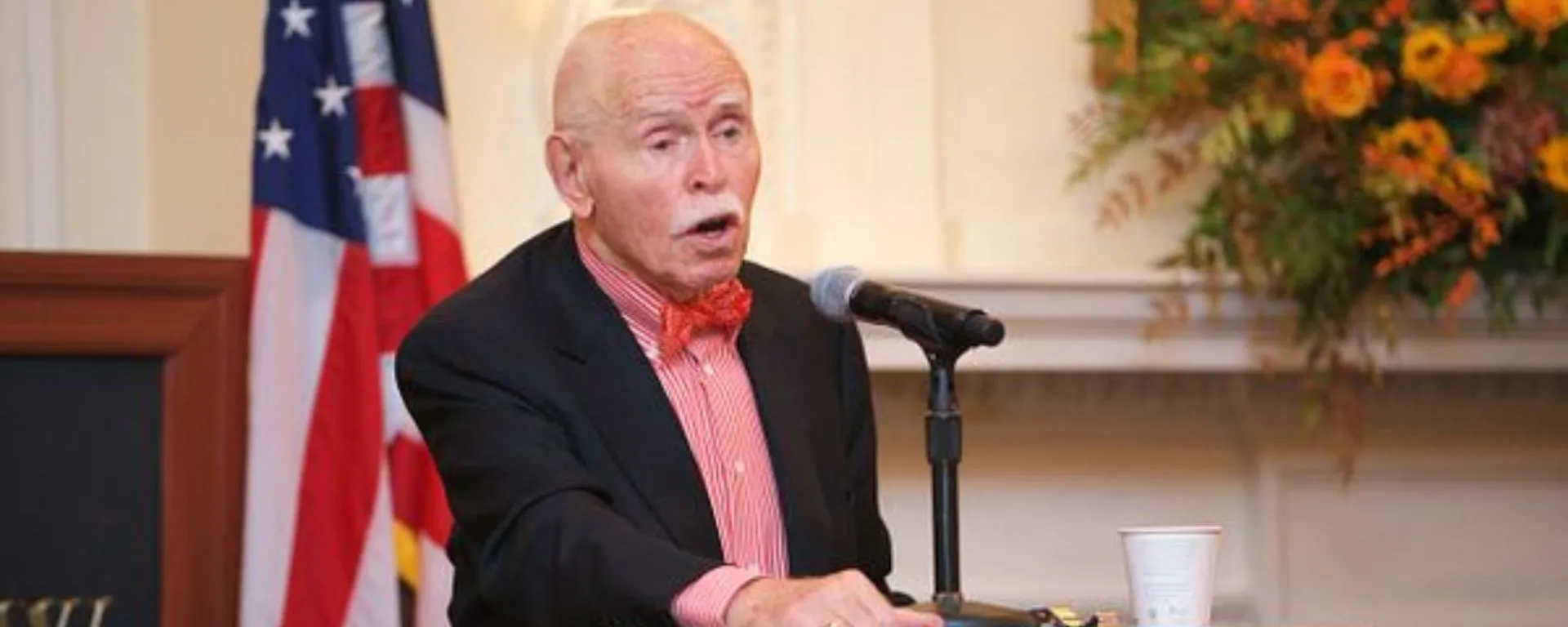 Jerome Cohen brindó conferencia sobre el triángulo Rusia-Ucrania-China Jerome Cohen en podio, al micrófono, con bandera estadounidense de fondo