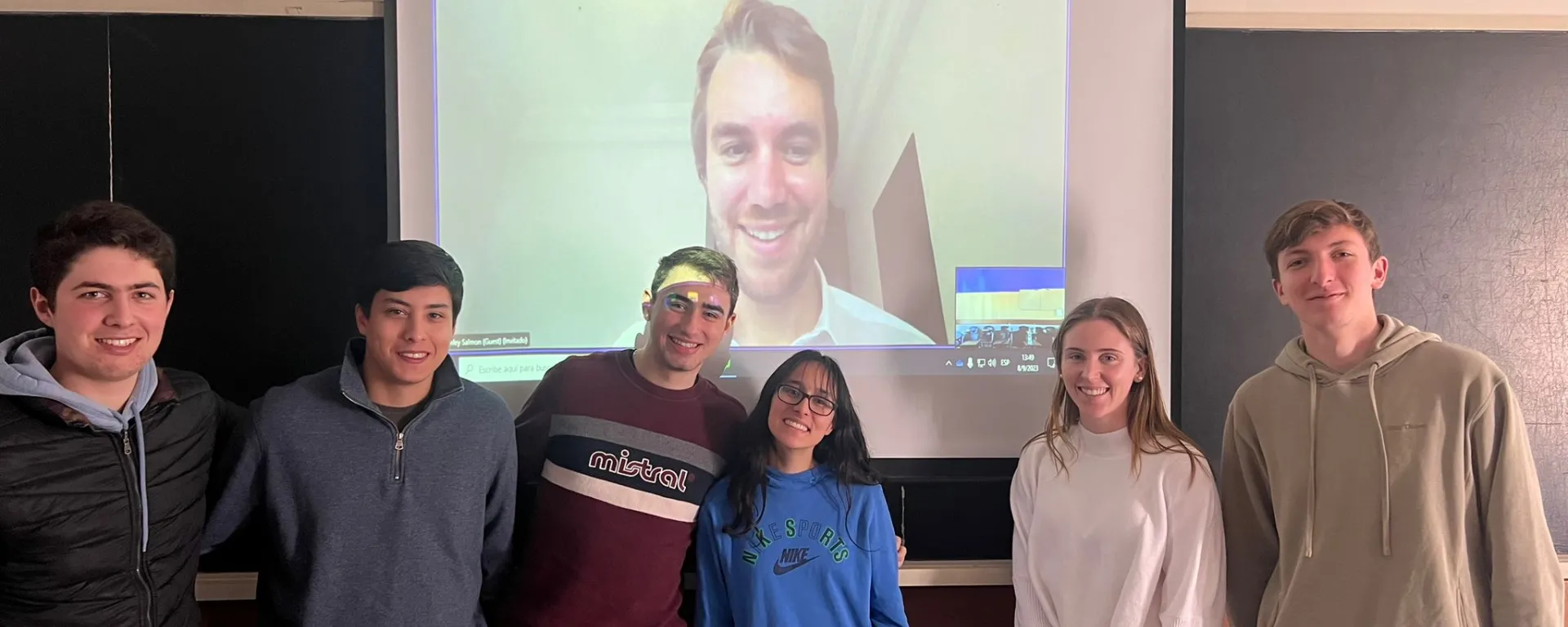 Los estudiantes que viajarán a Ghana. En la pantalla, el corresponsal de The Economist para África. Seis estudiantes universitarios miran a cámara. Detrás se ve una pantalla con un hombre.